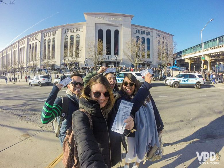 Foto da Bia com os amigos segurando os ingressos em frente ao estádio dos Yankees 