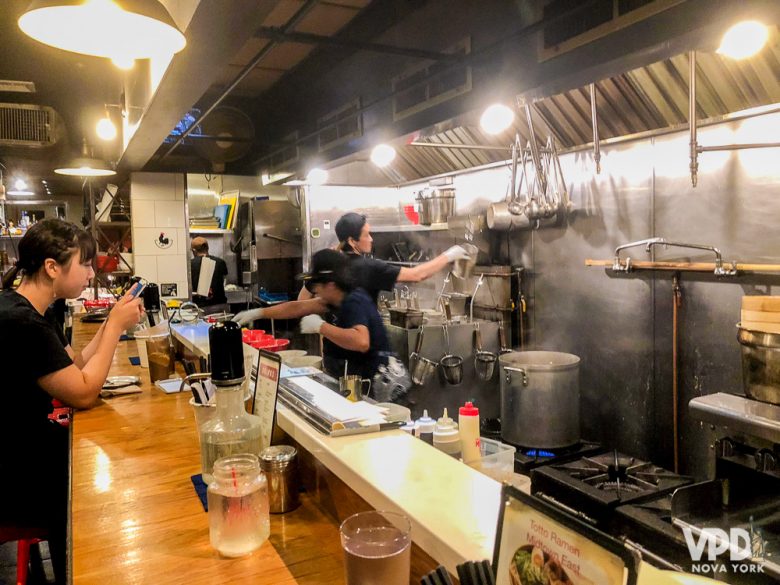 Foto do balcão do restaurante, com os chefs preparando a comida atrás - também uma opção para sentar em dupla ou sozinho.