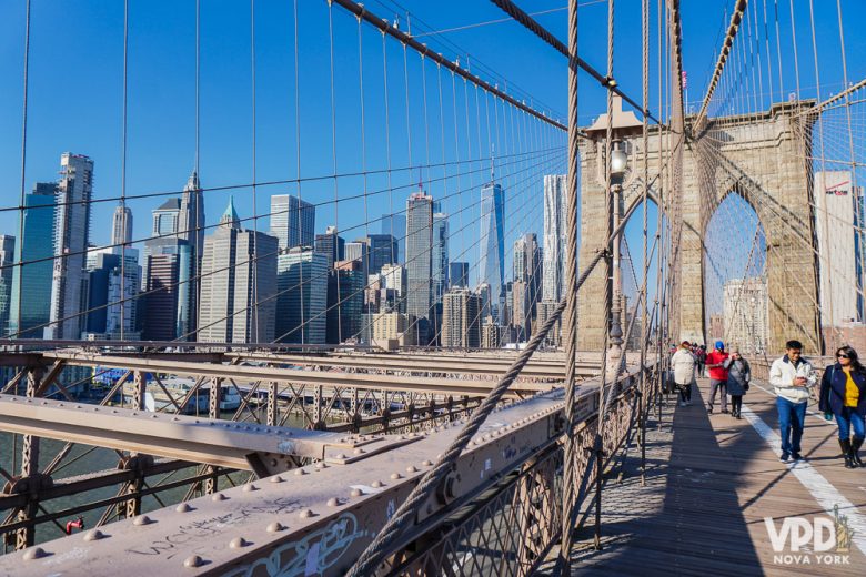 Foto da Brooklyn Bridge, com o céu azul e prédios ao fundo. 