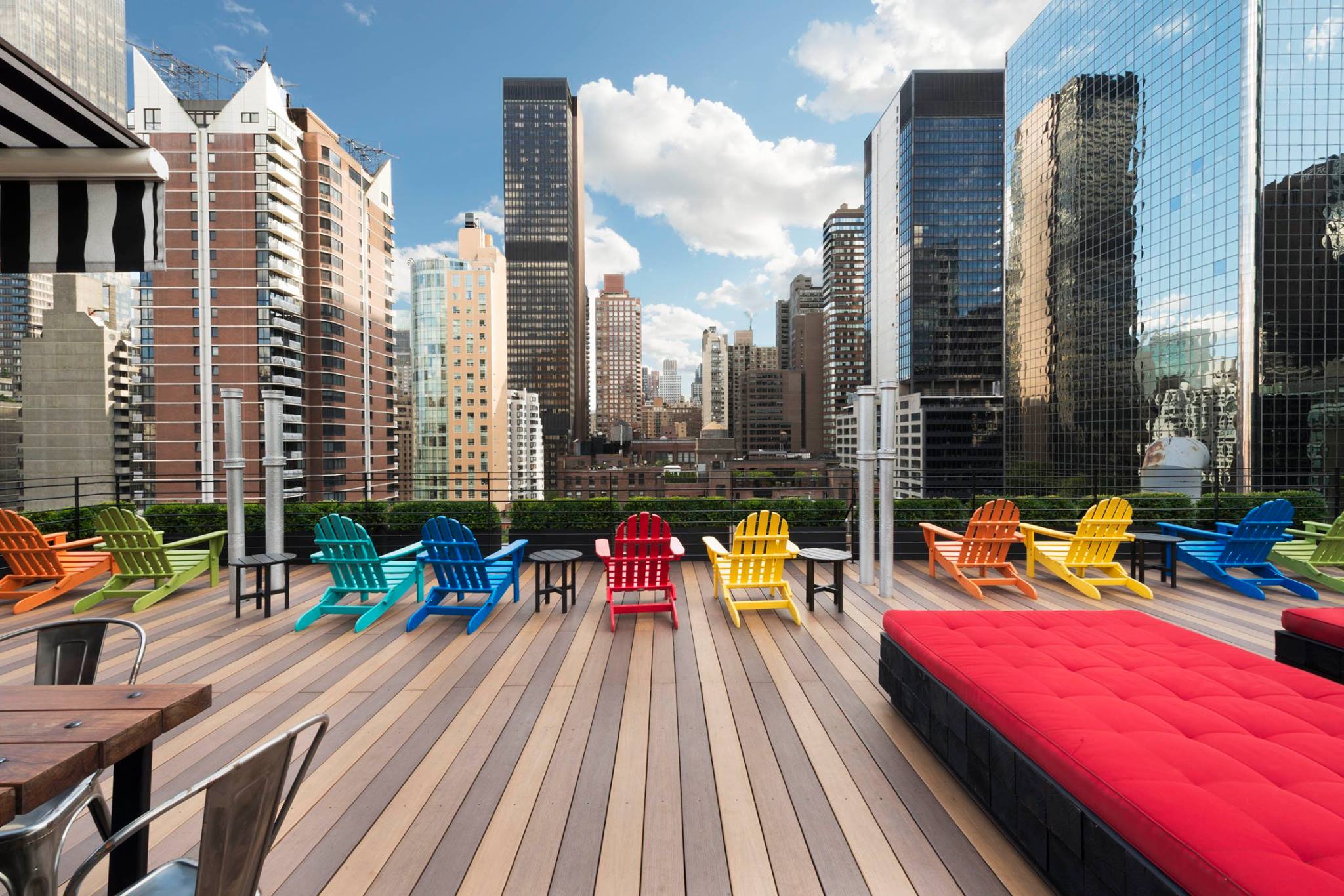 Foto da varandinha no telhado do hotel The Pod 51 em Nova York, com cadeiras coloridas para tomar sol, um sofazinho vermelho e os prédios da cidade ao fundo