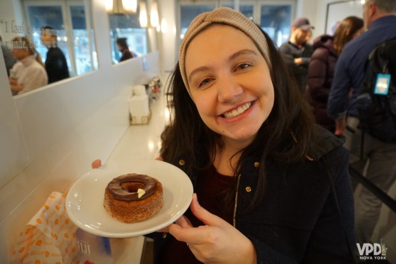 Foto da Renata segurando um prato com um cronut e sorrindo - uma das muitas opções de comida boa em Nova York. 
