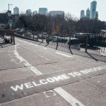 Foto de um escrito que diz "Welcome to Brooklyn" no chão da rua, com os prédios e o céu de Nova York ao fundo