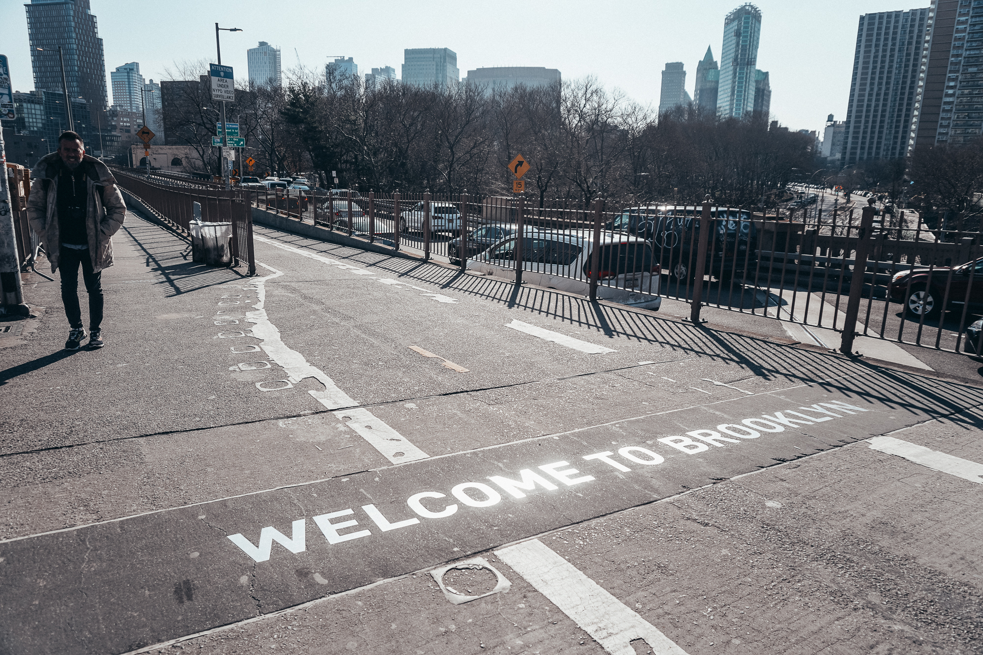 Foto de um escrito que diz "Welcome to Brooklyn" no chão da rua, com os prédios e o céu de Nova York ao fundo