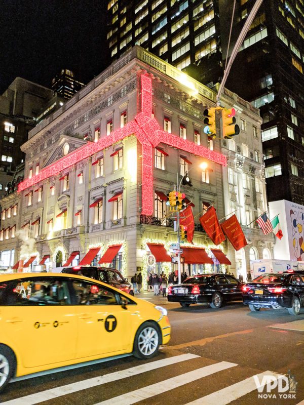 Foto de uma loja com um laço de presente feito de luzinhas de Natal vermelhas e um taxi amarelo passando em frente