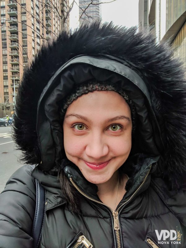 A imagem mostra a Renata com um casaco grande de inverno com capuz e gorro. Nova York em janeiro. 