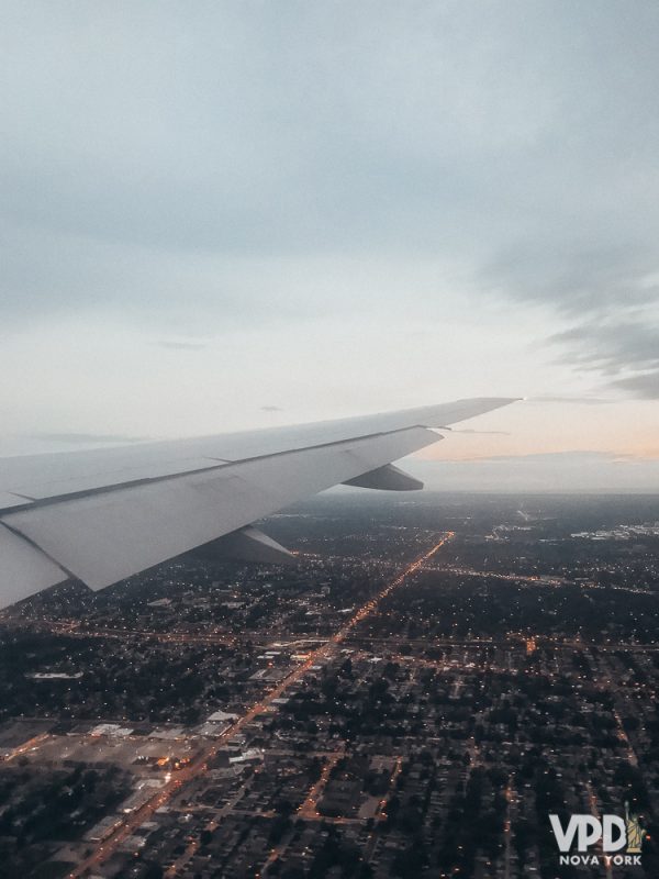 É importante entender o custo-benefício de cada plano de seguro viagem. Foto da asa de um avião sobrevoando a cidade de Nova York, com um céu cinza-claro ao fundo 