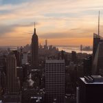 Foto da cidade de Nova York no pôr do sol, com os prédios escuros e o céu azul, rosa e laranja
