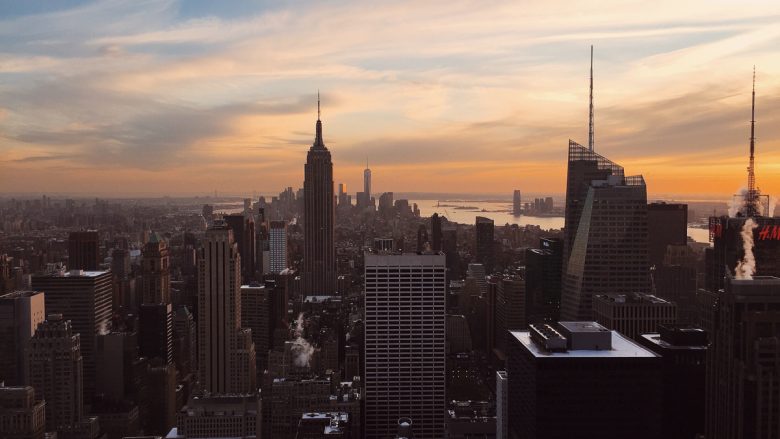 Foto da cidade de Nova York no pôr do sol, com os prédios escuros e o céu azul, rosa e laranja