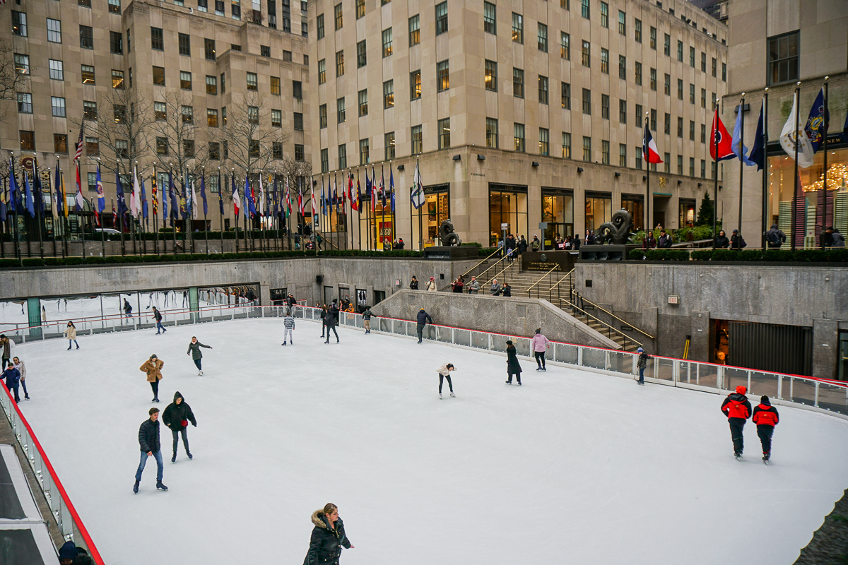 Imagem da pista de patinação do Rockfeller Center. Há pessoas patinando e bandeiras e prédios ao redor.