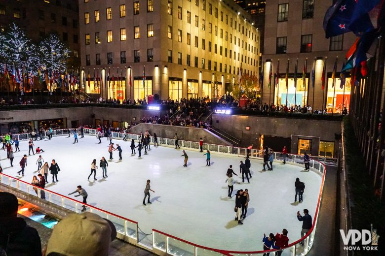 Imagem da pista de patinação do Rockefeller Center em fevereiro. Está de noite e várias pessoas patinam. Os prédios estão iluminados ao redor e muitas pessoas assistem os patinadores.