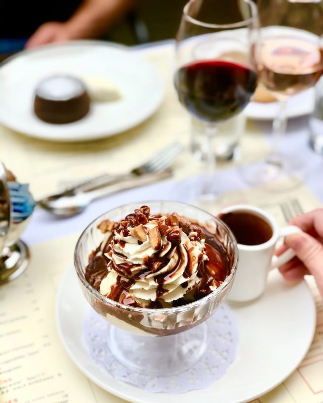 O Harry's Fudge Sundae do Cafe Luxembourg. Um potinho de vidro com sorvete, calda de chocolate, chantilly e amêndoas. No fundo, há talheres e taças de vinho.