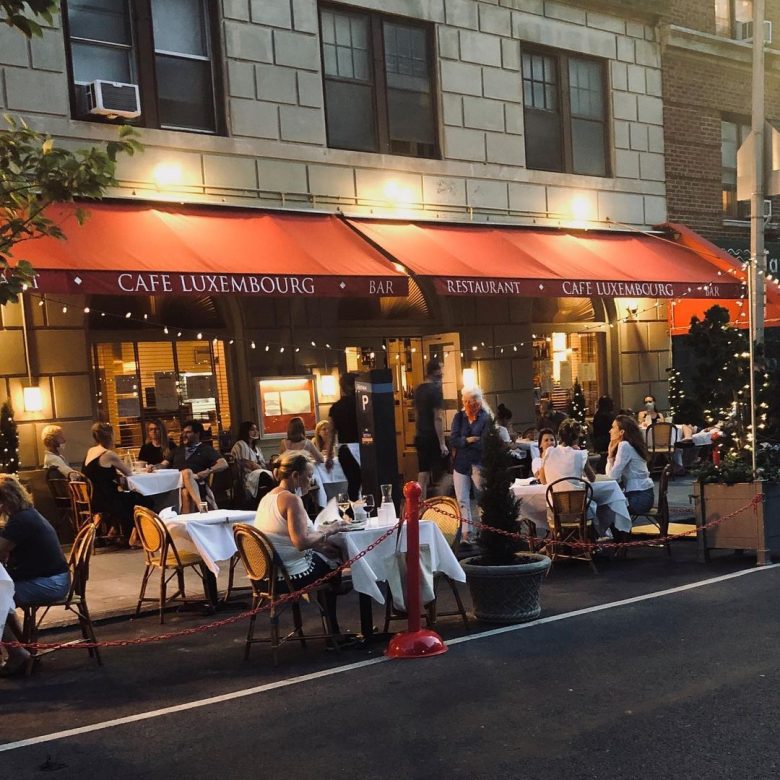 Imagem da fachada do Cafe Luxembourg. As mesas estão espalhadas na calçada e há fios de luzinhas.