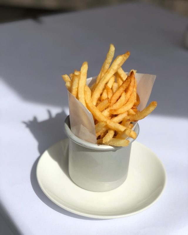 Imagem de uma porção de batatas fritas. Elas estão dentro de um potinho branco que está apoiado em um pratinho branco e em uma mesa branca. 