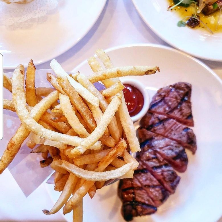 Imagem de uma porção de batatas fritas, um potinho com ketchup e um filé de carne vermelha em um prato branco. O steak frites é um dos pratos do Cafe Luxembourg.