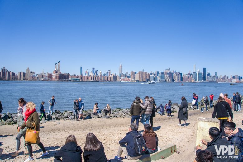 Imagem de Nova York em março. Várias pessoas estão sentadas nas margens do rio, e o Skyline da cidade está ao fundo. 