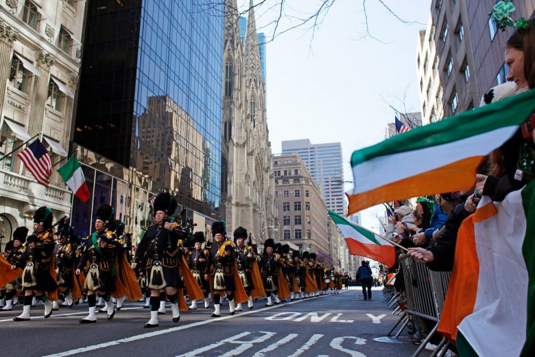 Imagem de uma parada de St Patrick's Day que acontece em março. Algumas pessoas marcham na rua e outras assistem segurando bandeiras da Irlanda nas laterais.