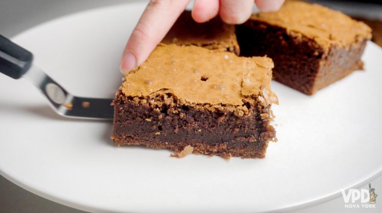 Imagem de um quadradinho de brownie de chocolate sendo colocado no prato. Ao fundo, há outros dois pedaços de brownie.