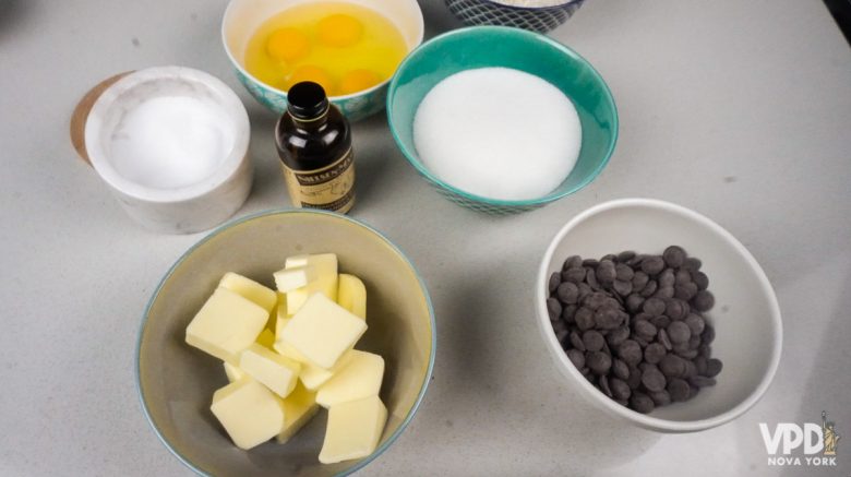 Foto com os ingredientes do brownie. Há potinhos com ovos, sal, açúcar, manteiga, baunilha e gotas de chocolate.