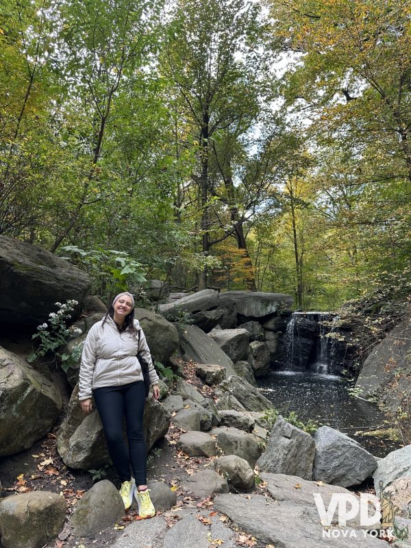 Foto da Re de pé em uma área de lago com uma pequena cachoeira. Ela usa um casaco e há várias árvores ao redor.