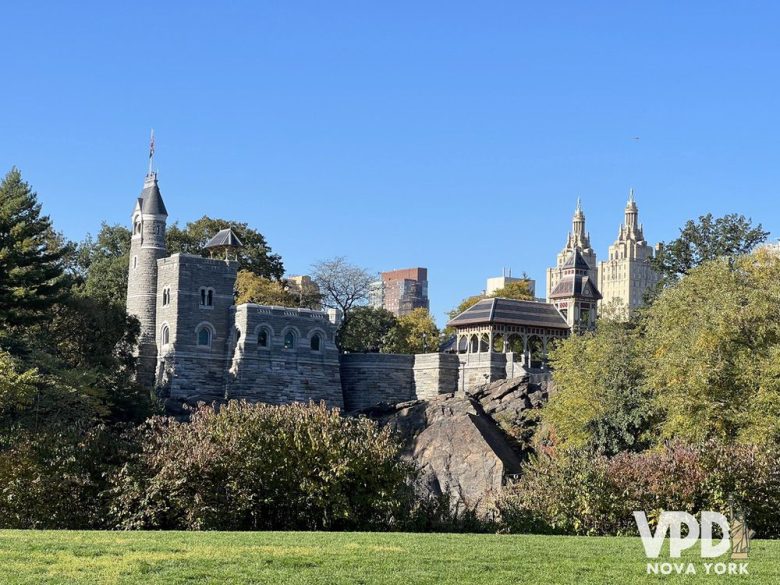 Foto do castelo no meio do central park. Há rochas ao redor, e ao fundo dá pra ver alguns prédios mais altos.