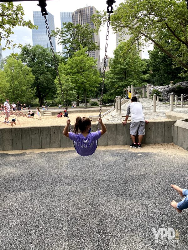 Foto de uma criança em uma balança de um dos parquinhos do Central Park. Ao fundo, dá pra ver várias crianças brincando.