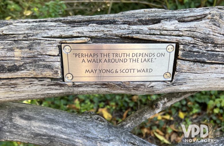 Foto de uma placa em um banco de madeira do parque. Ele traz uma frase que diz: "talvez a verdade dependa de um passeio ao redor do lago". Os nomes abaixo da frase são May Yong e Scott Ward.