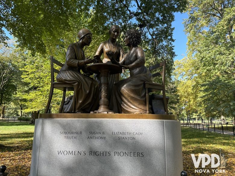 Foto de uma escultura que representa três ativistas sentadas em uma mesa. Os nomes delas estão escritos abaixo, e há várias árvores ao redor.