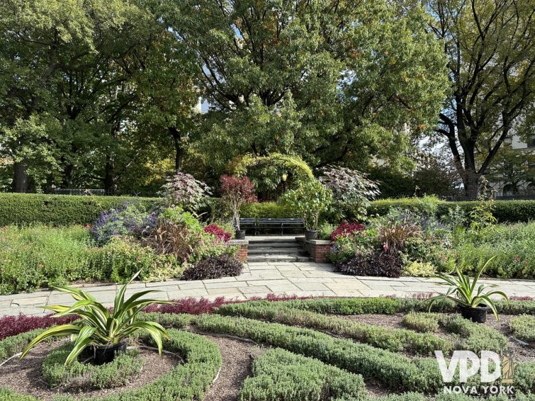 foto de um jardim com muitas plantas verdes, vermelhas e roxas. há um arco feito com plantas e árvores ao fundo.