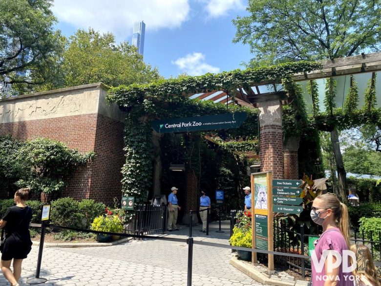 Foto da entrada do Central Park Zoo. Ele tem várias plantas e pessoas andando para entrar.