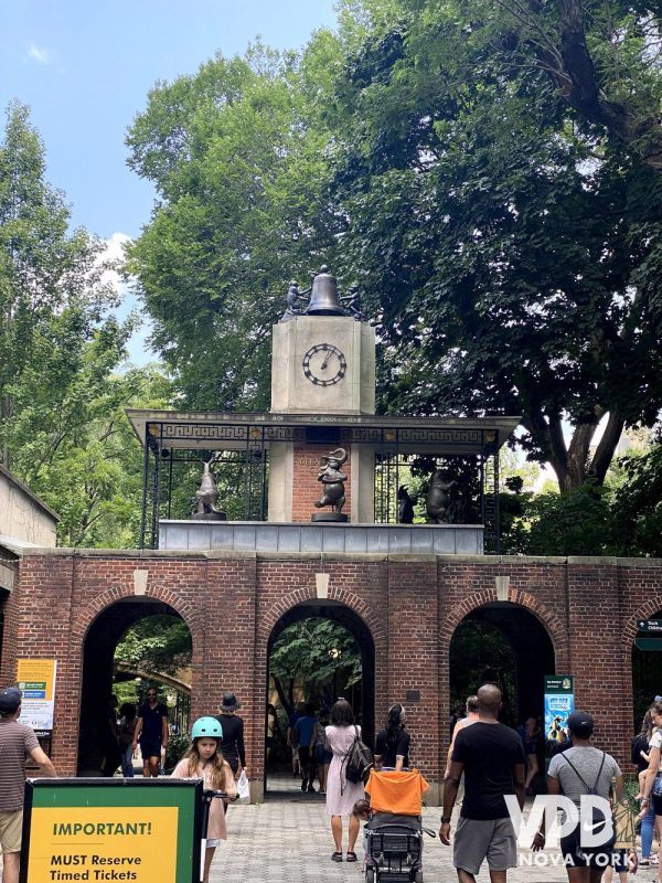 Foto do Delacorte Clock, um relógio no alto de uma torre com esculturas ao redor. Abaixo, várias pessoas cruzam o parque.